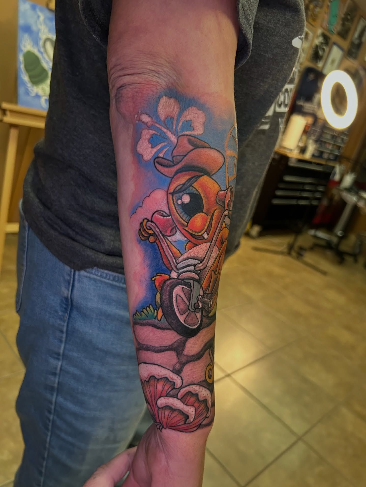 Tattoo of Deadpool riding a motorcycle, wearing a cowboy hat. Colorful design on forearm with blue, pink, and orange hues.