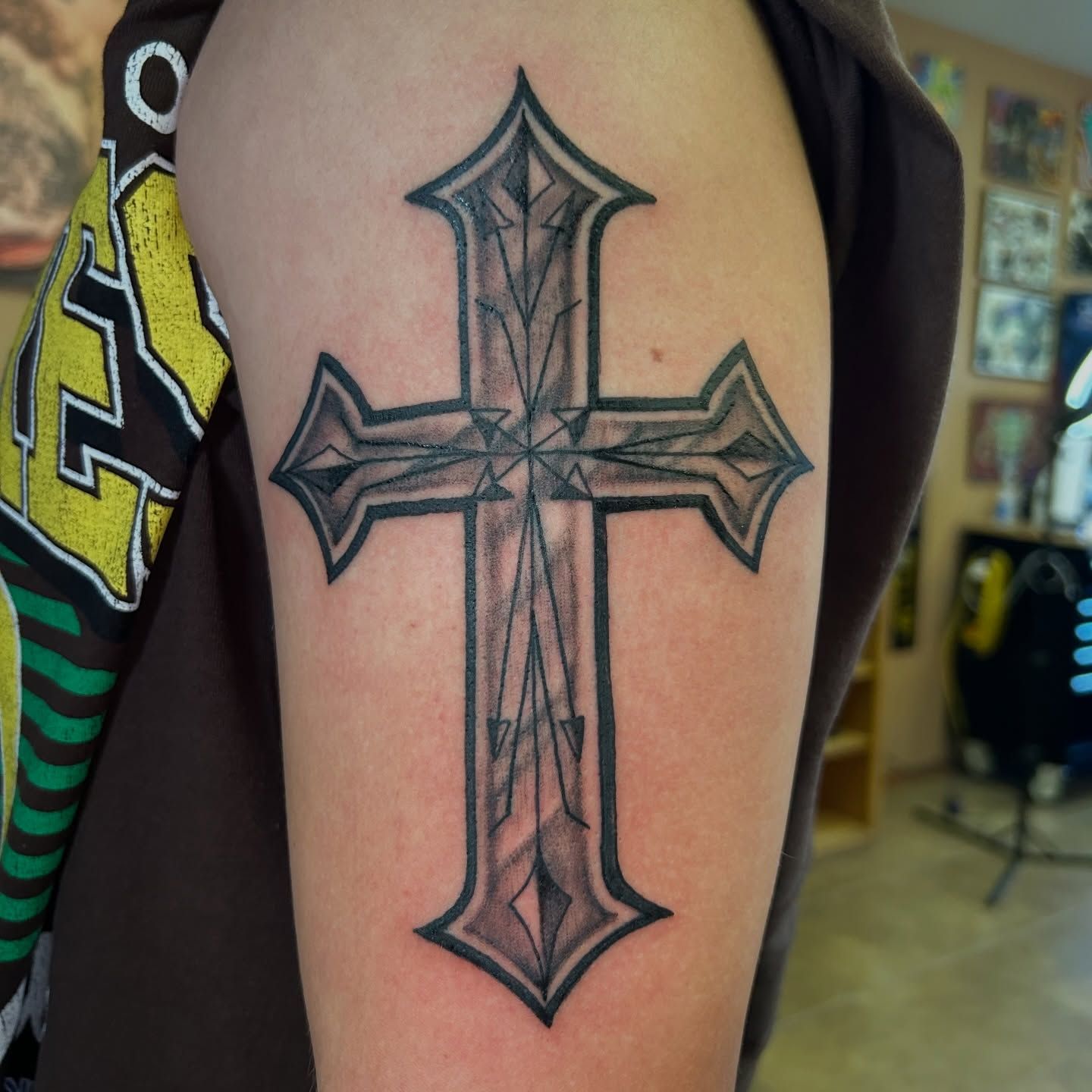 Tattoo of a black and gray cross on a person's upper arm, with ornamental details.