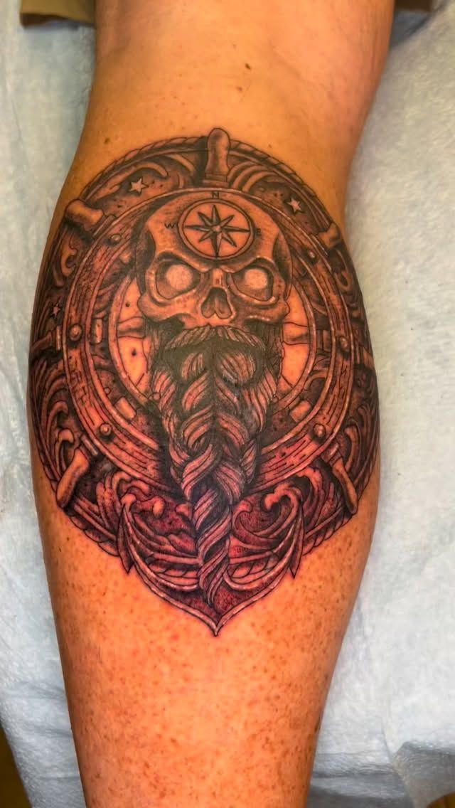 Tattoo of a skull-faced dwarf with a braided beard, inside a circular, ornate design on a leg.