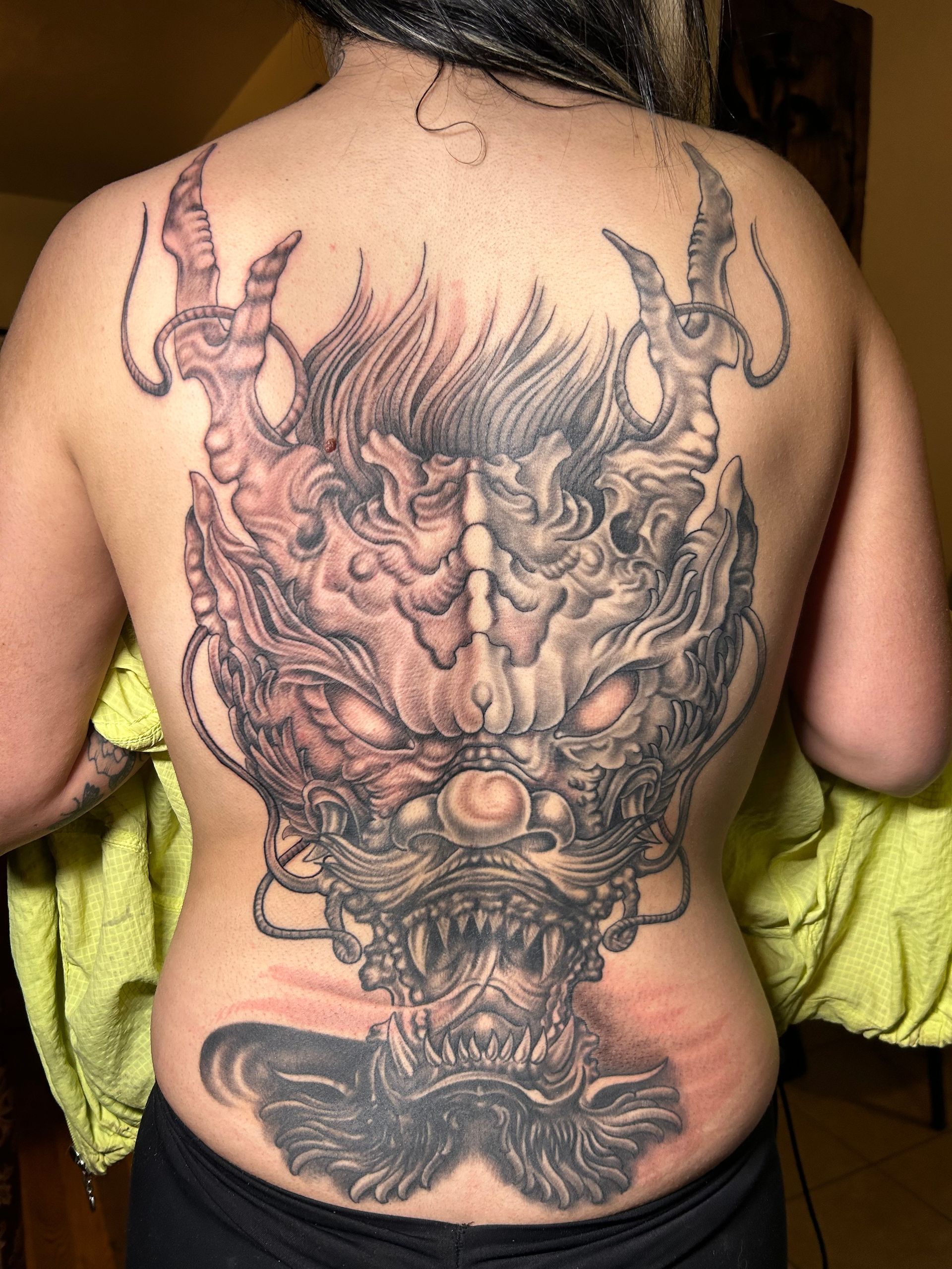 Tattoo of a detailed, gray and red oni mask covers a person's back, with a smaller design on their lower back.