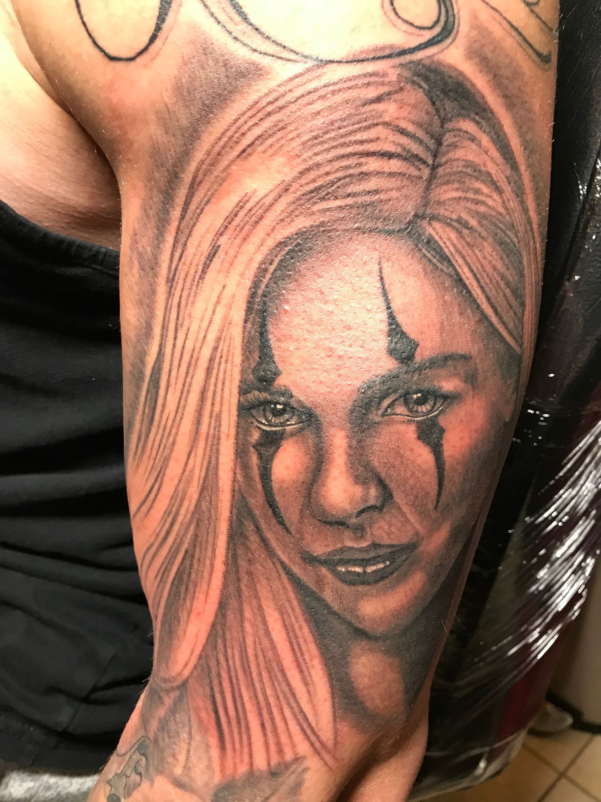 Black and gray tattoo of a person with blonde hair and clown-like face paint on an arm.