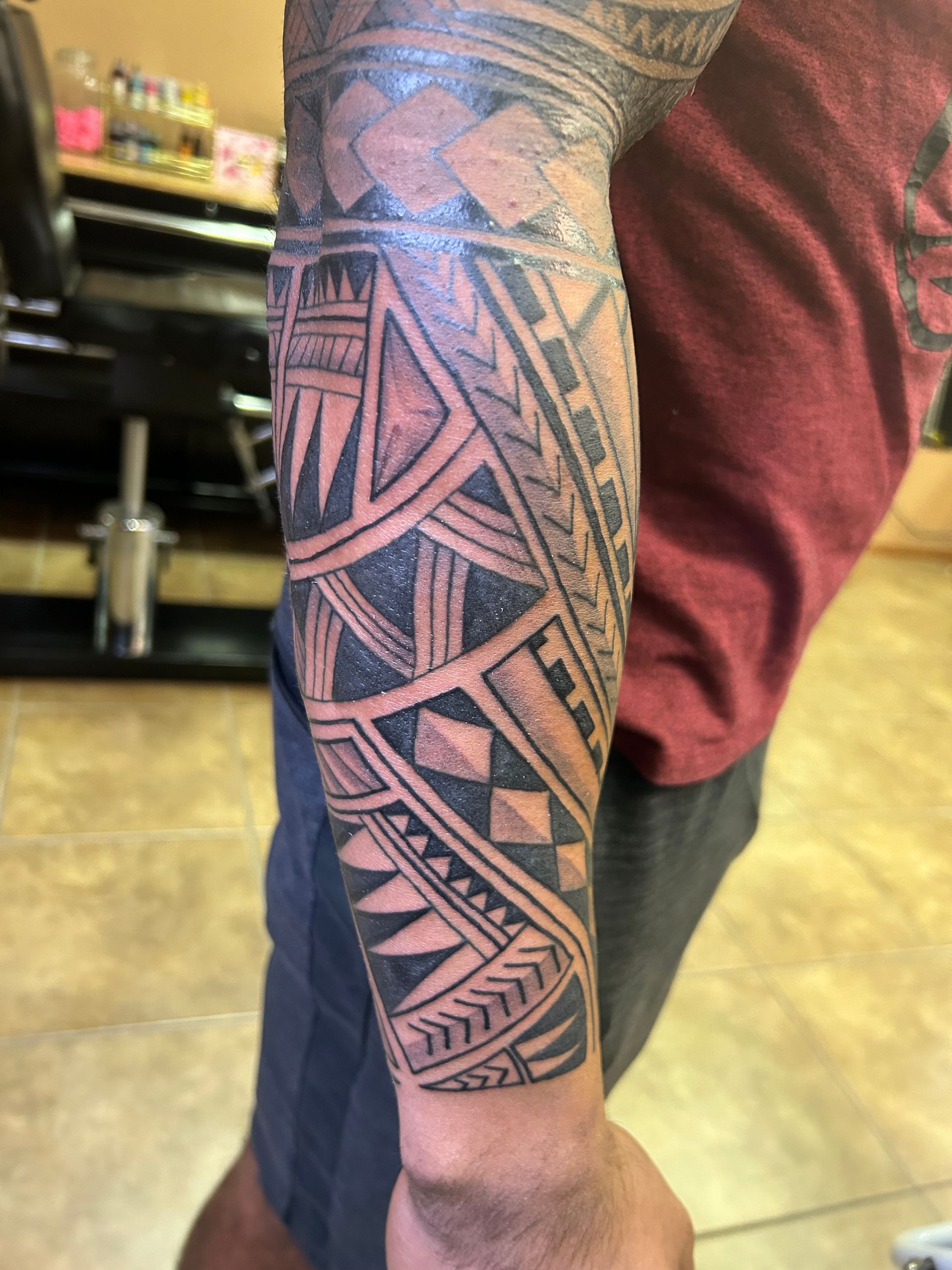 Arm with Polynesian tribal tattoo. Dark ink design covering the forearm. Person wearing a red shirt and shorts.