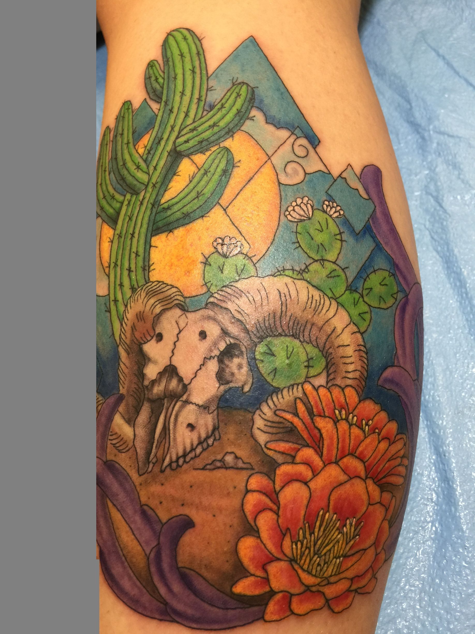 Tattoo of a desert scene with a ram, cacti, orange flowers, and mountains in blue and yellow.