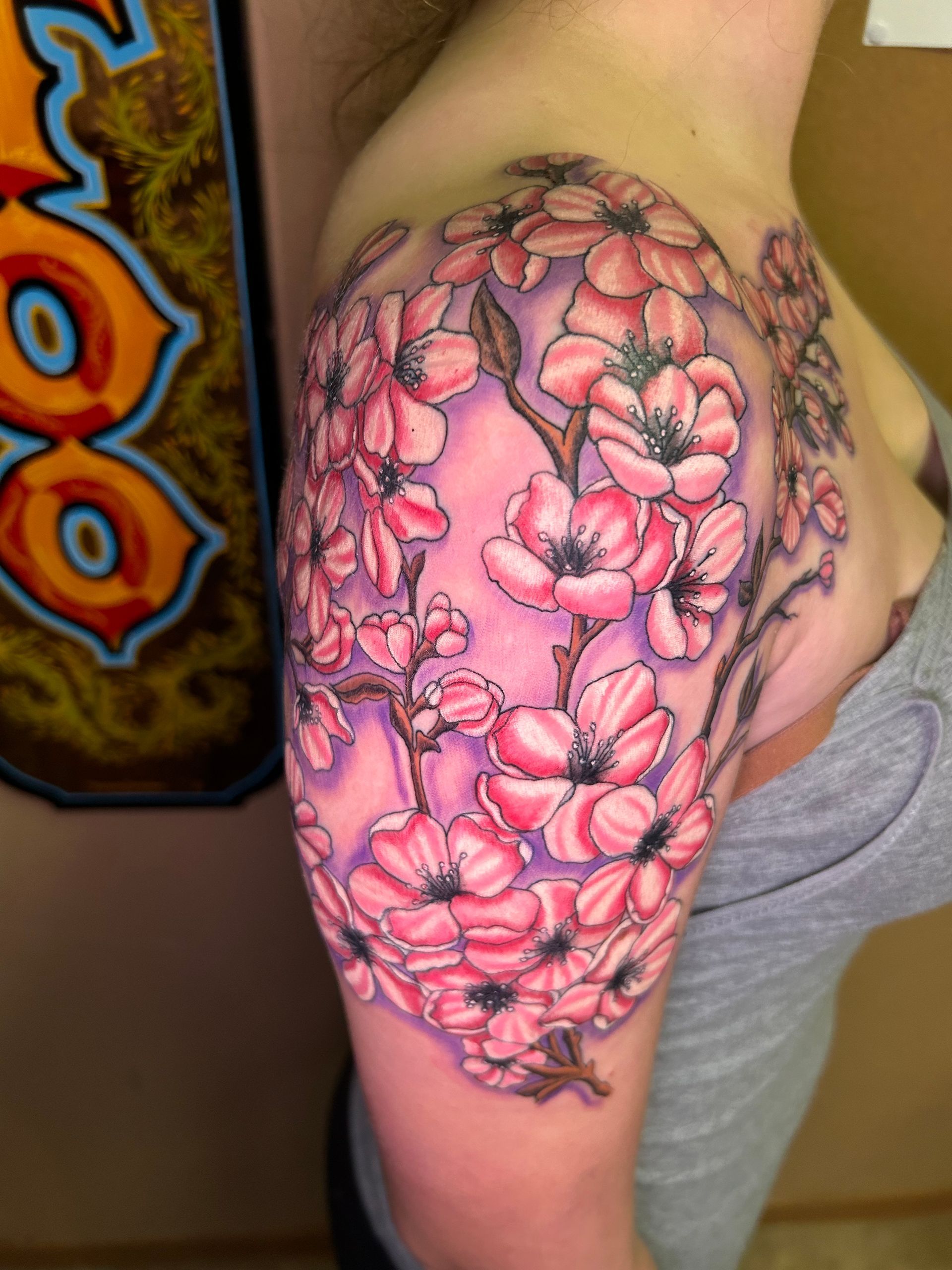Tattoo of pink cherry blossoms on a person's shoulder and upper arm with a purple background.