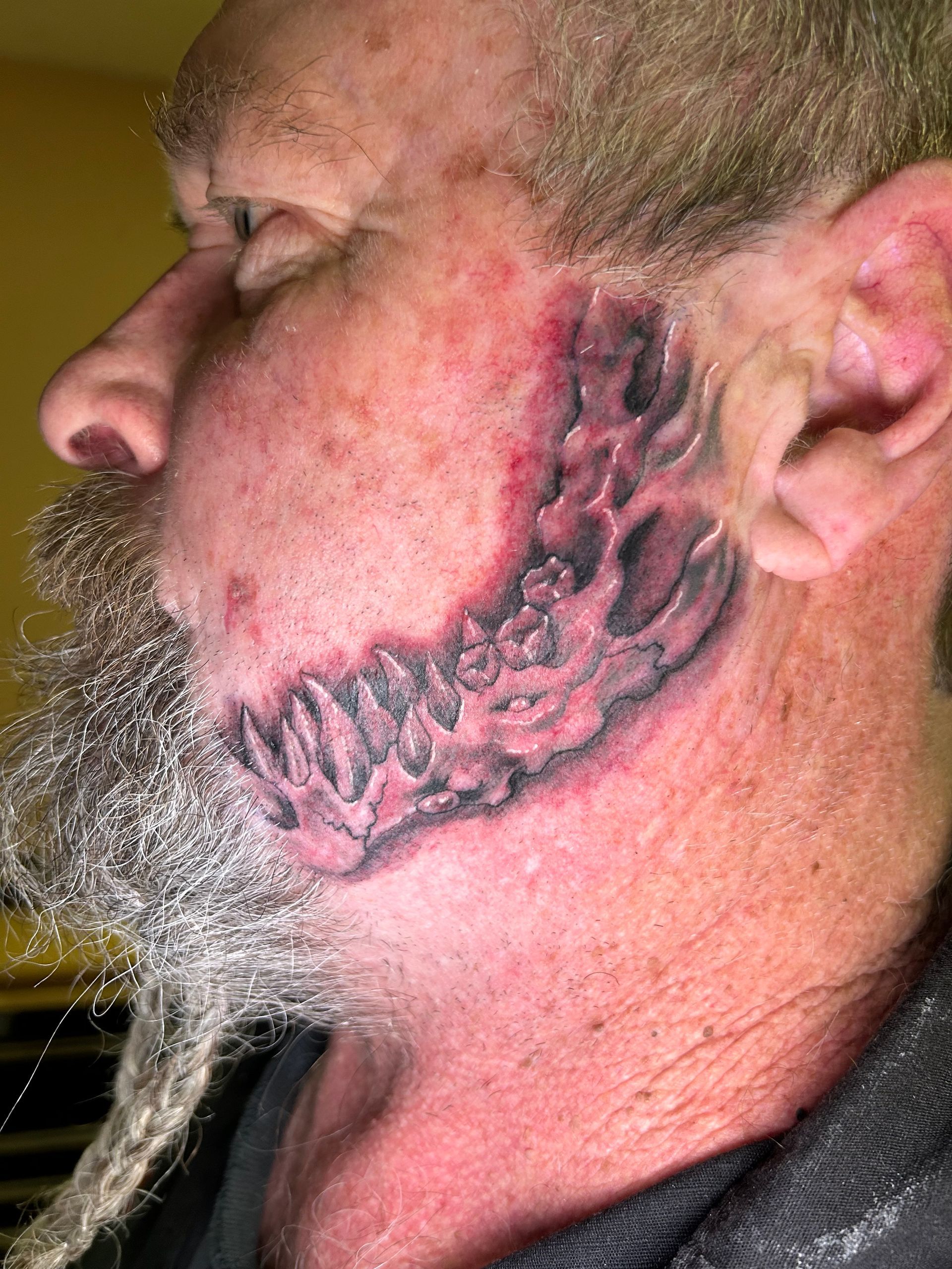 Man's face with a dark, detailed tattoo on the cheek and jawline, along with a grey beard.