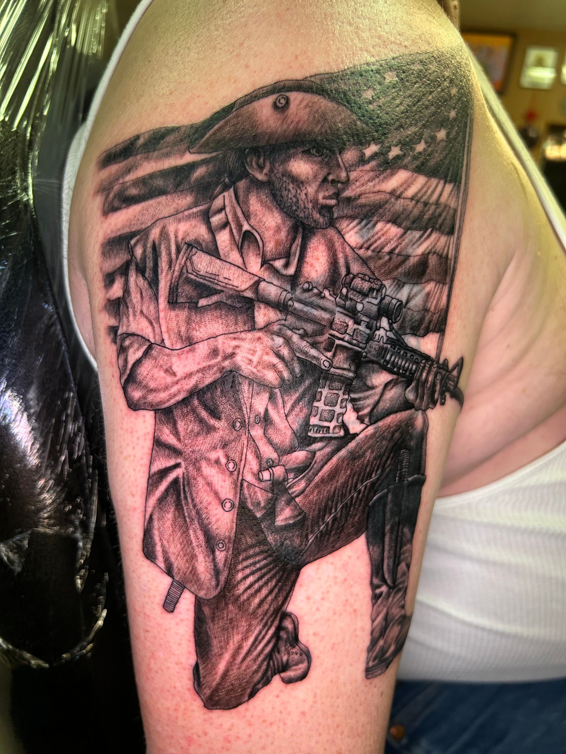 Tattoo of a soldier kneeling, holding a rifle, with an American flag background. Black and white.