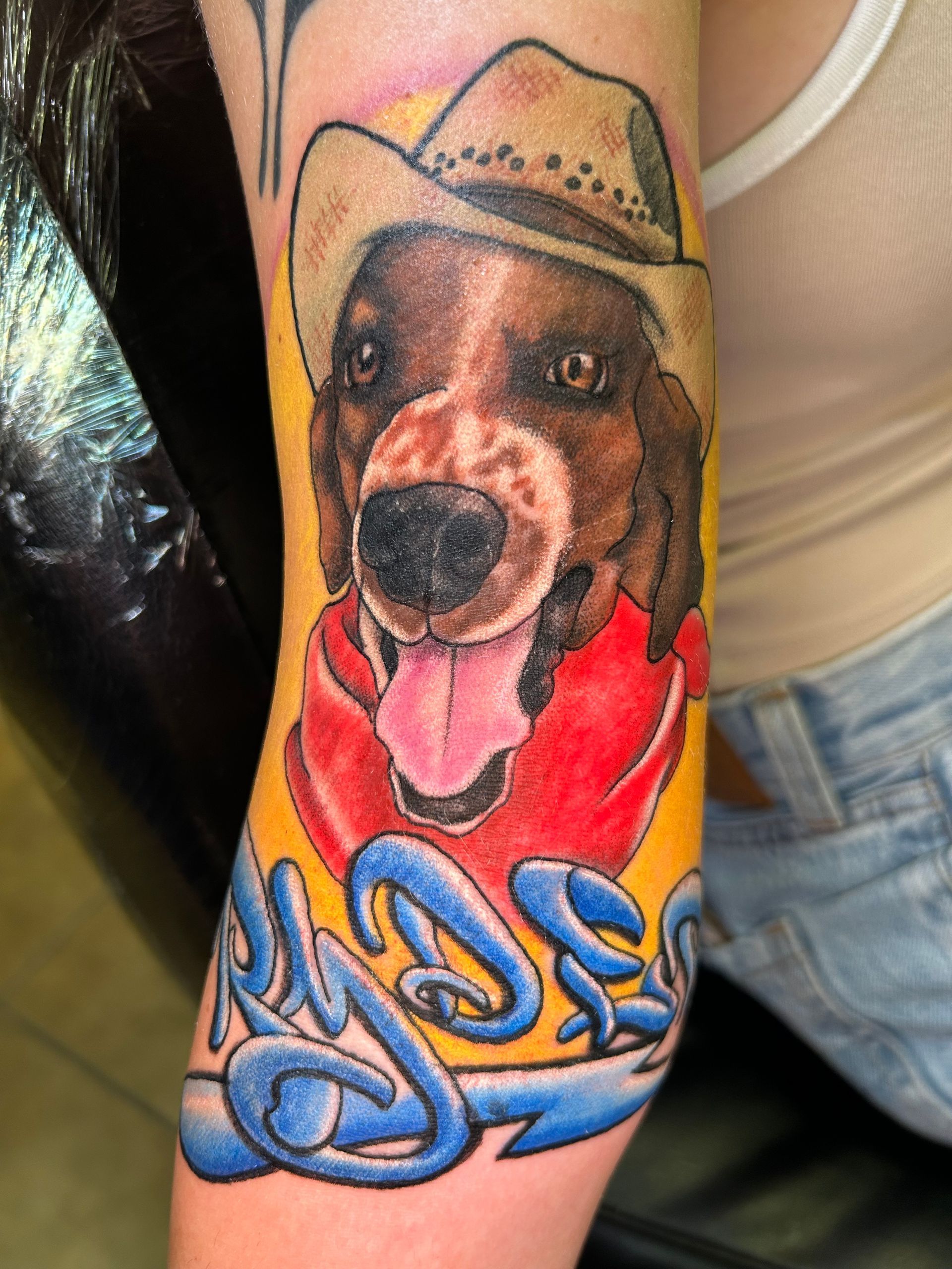 Tattoo of a dog wearing a cowboy hat and red jacket, with “My Dez” in blue lettering on a yellow background.