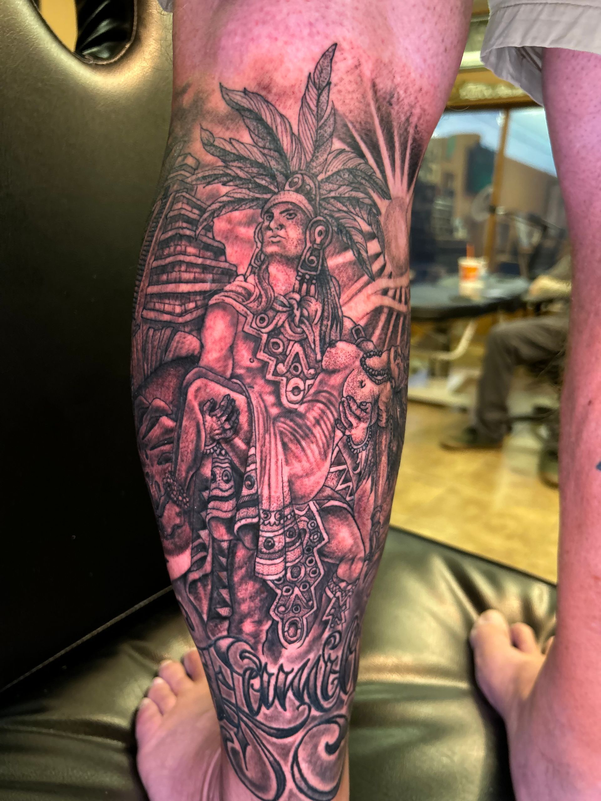 Detailed grayscale leg tattoo: Aztec deity with headdress, architecture, and rising sun.