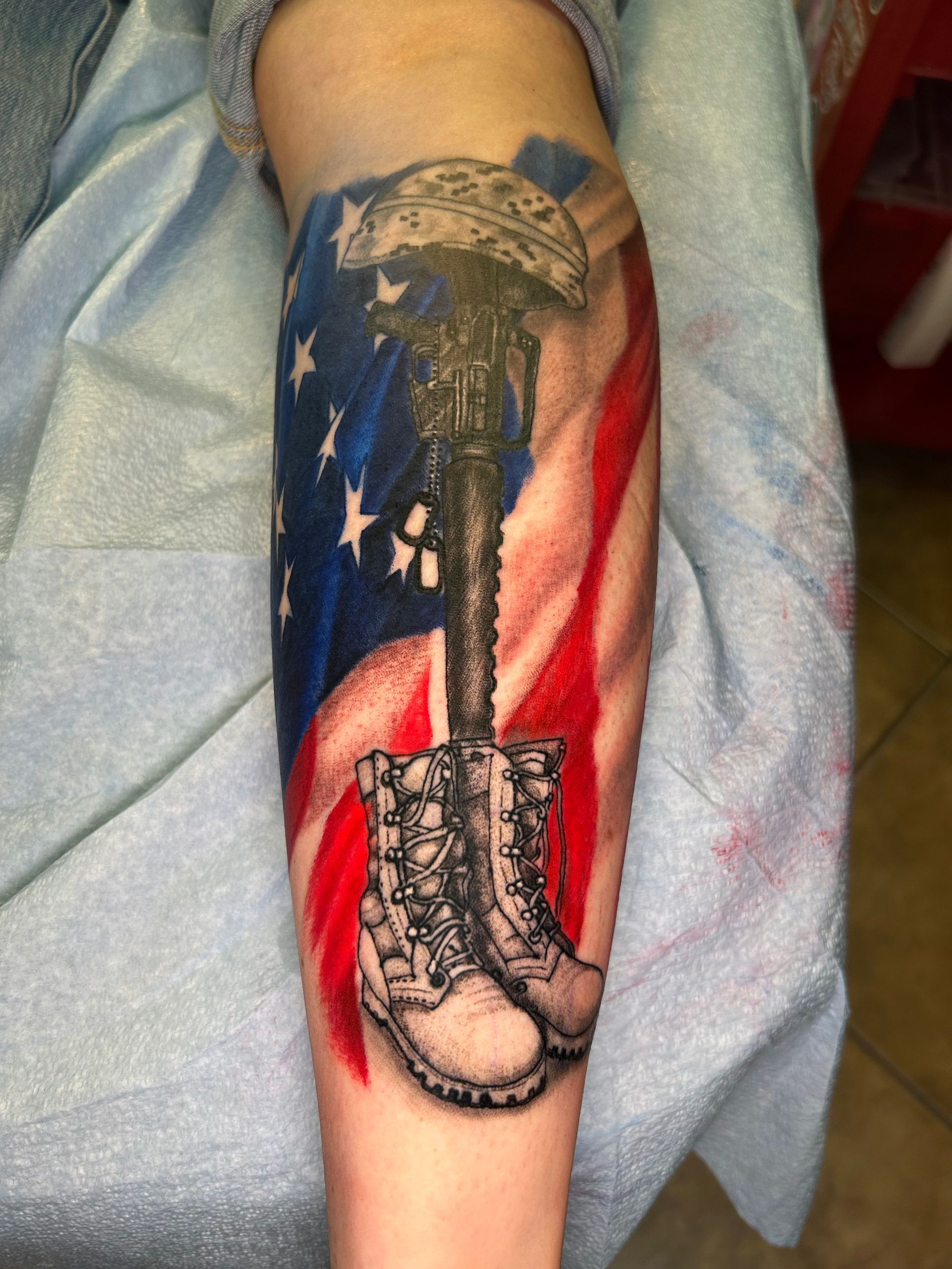 Tattoo of a soldier's fallen boots and rifle, with an American flag background on an arm.