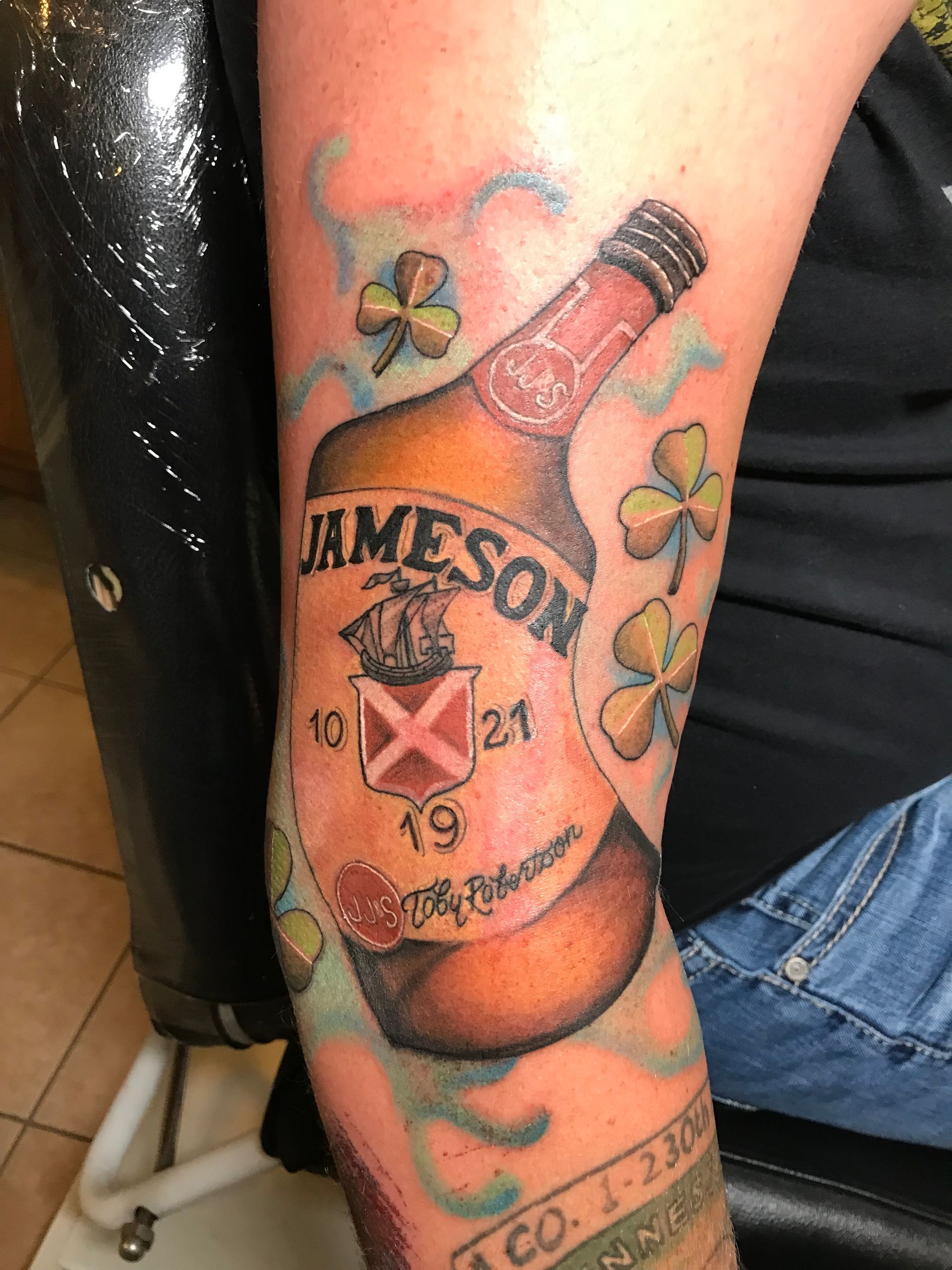 Tattoo of a Jameson whiskey bottle with shamrocks. Arm is visible; the tattoo has green, brown, and red colors.