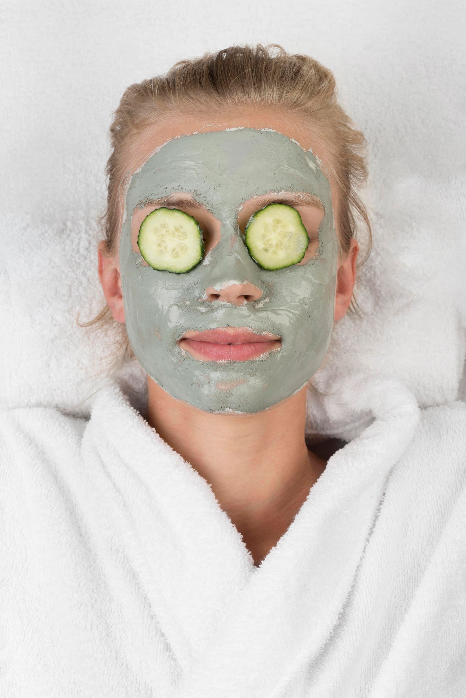 Person with face mask and cucumber slices on eyes, wearing a robe, lying on a white surface.