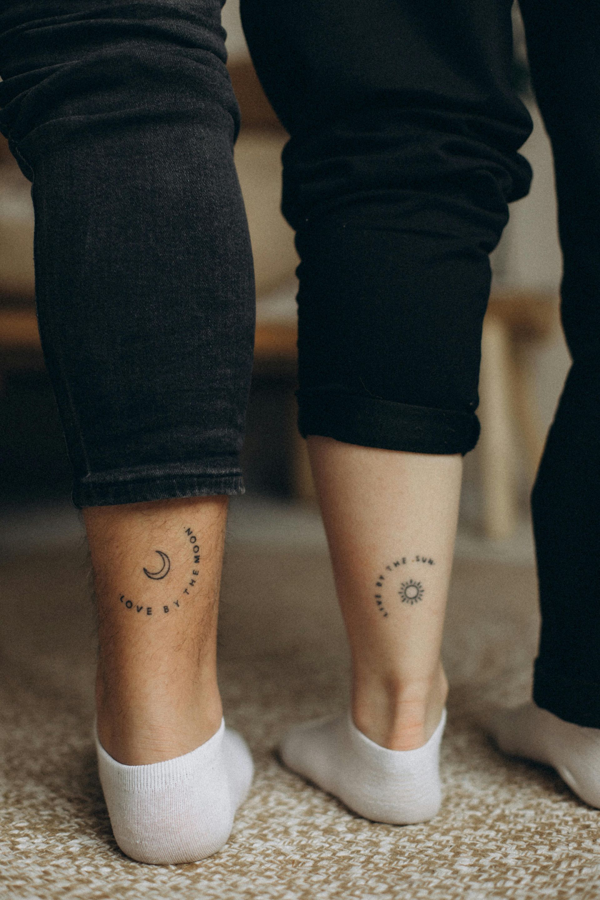 Two people's ankles with matching tattoos of the sun and moon; wearing black pants and white socks.