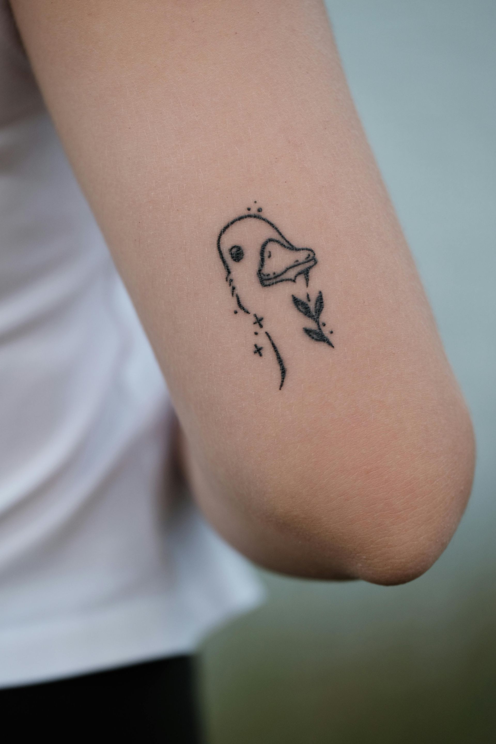 Black line art tattoo of a duck head on an arm, holding a leaf.