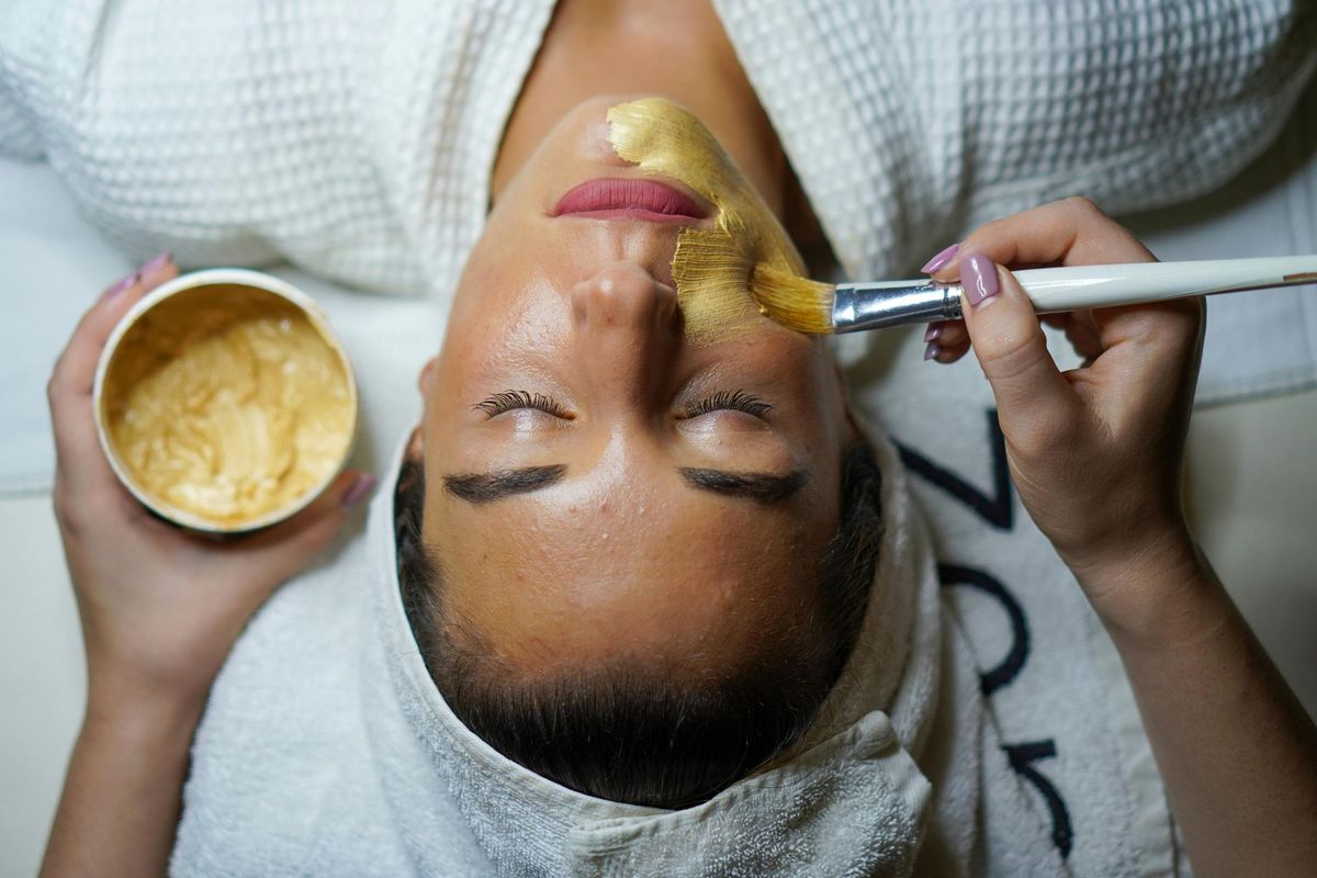 Woman receiving facial treatment with gold mask, lying on white towel, eyes closed.