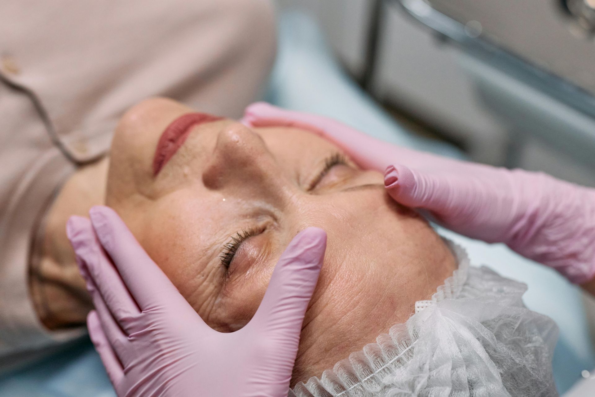 Woman getting a facial; hands in pink gloves on her face, eyes closed, in a medical setting.