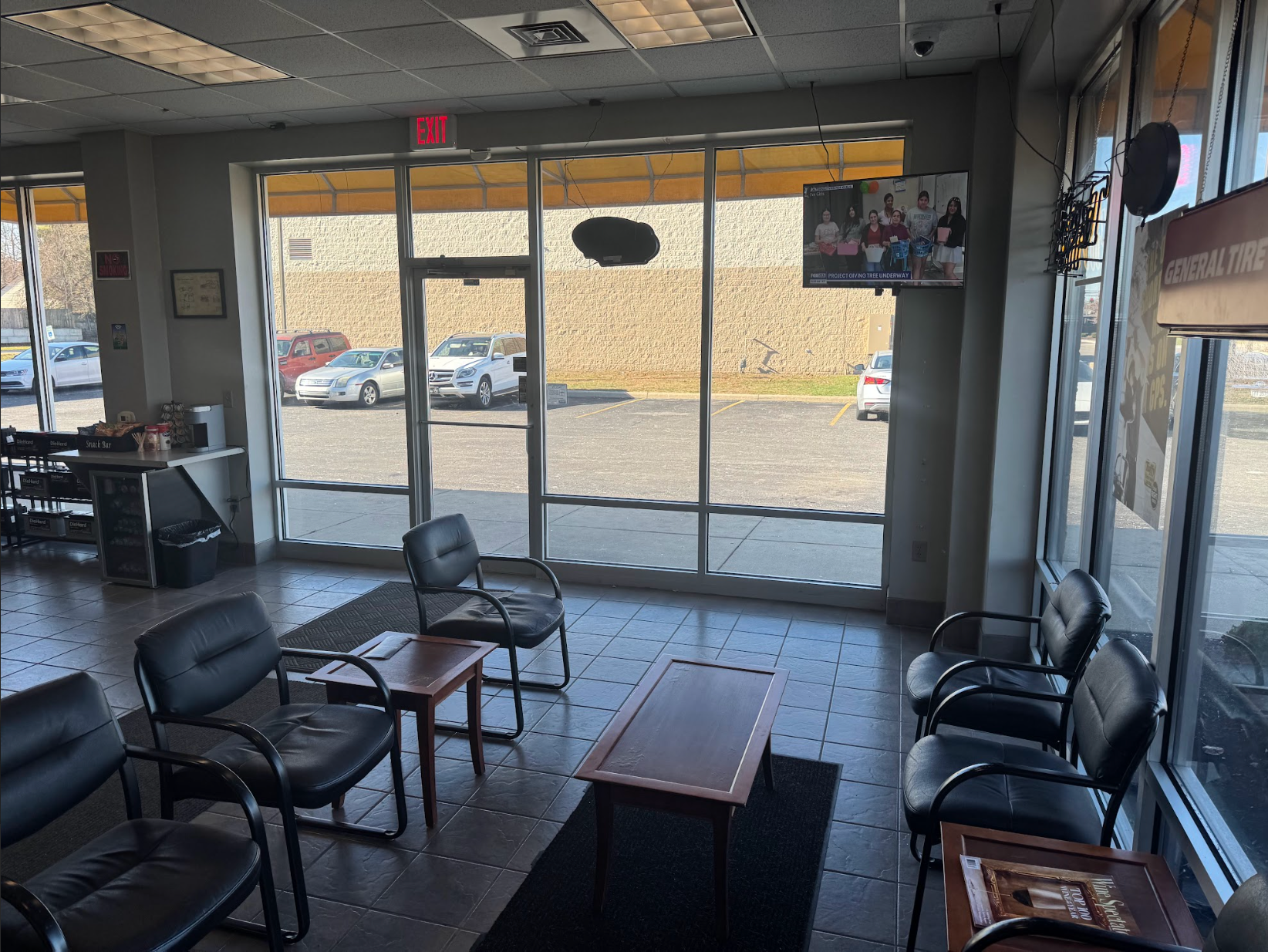 Waiting area with black chairs, tables, and a TV; large windows with cars outside.