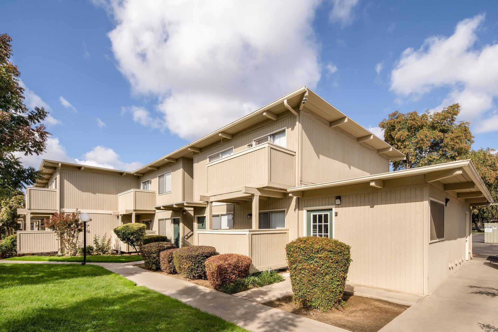 Two-story beige apartment building with balconies, green lawn, and bushes under a cloudy sky.