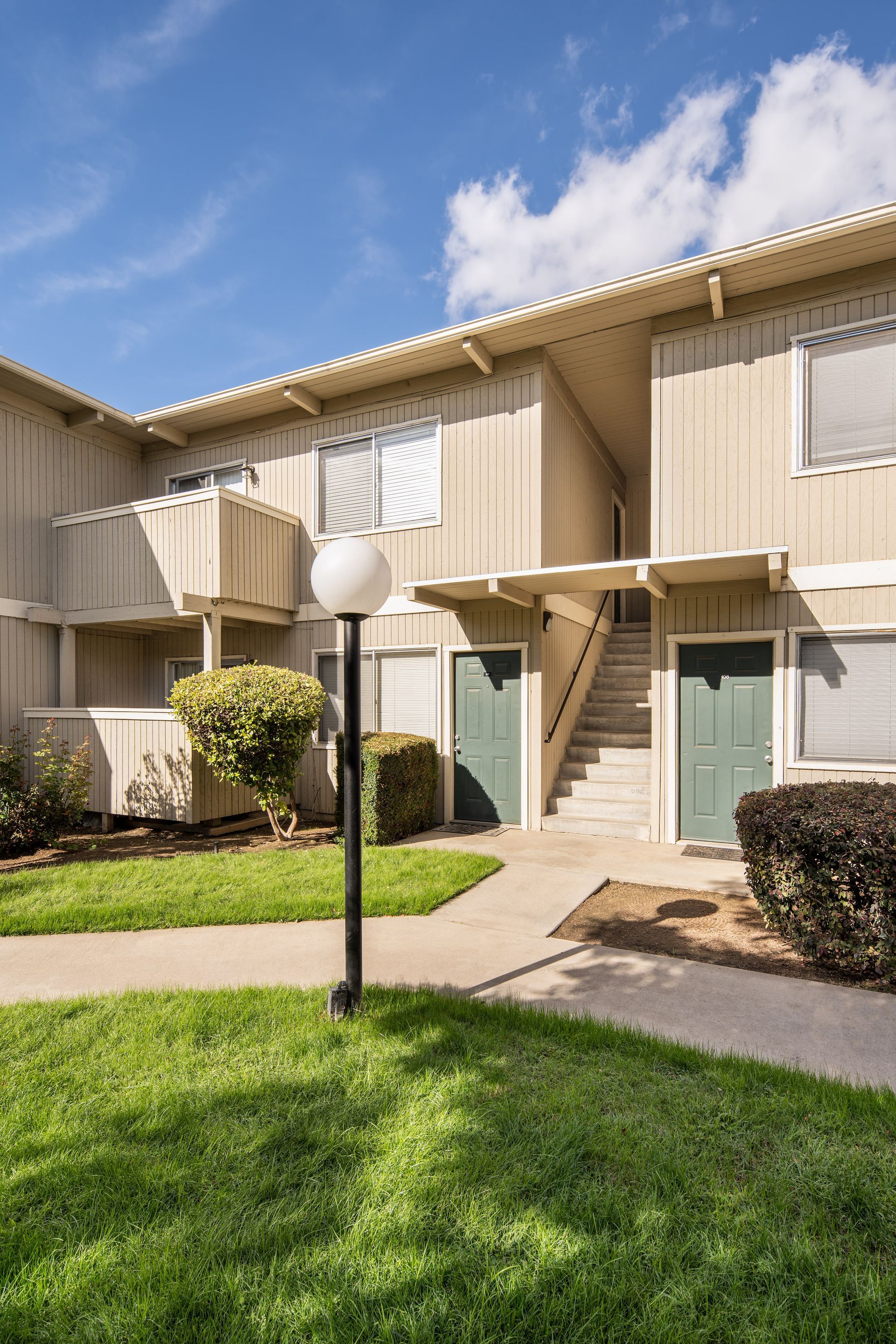 Two-story beige apartment building with green doors and landscaping, on a sunny day.