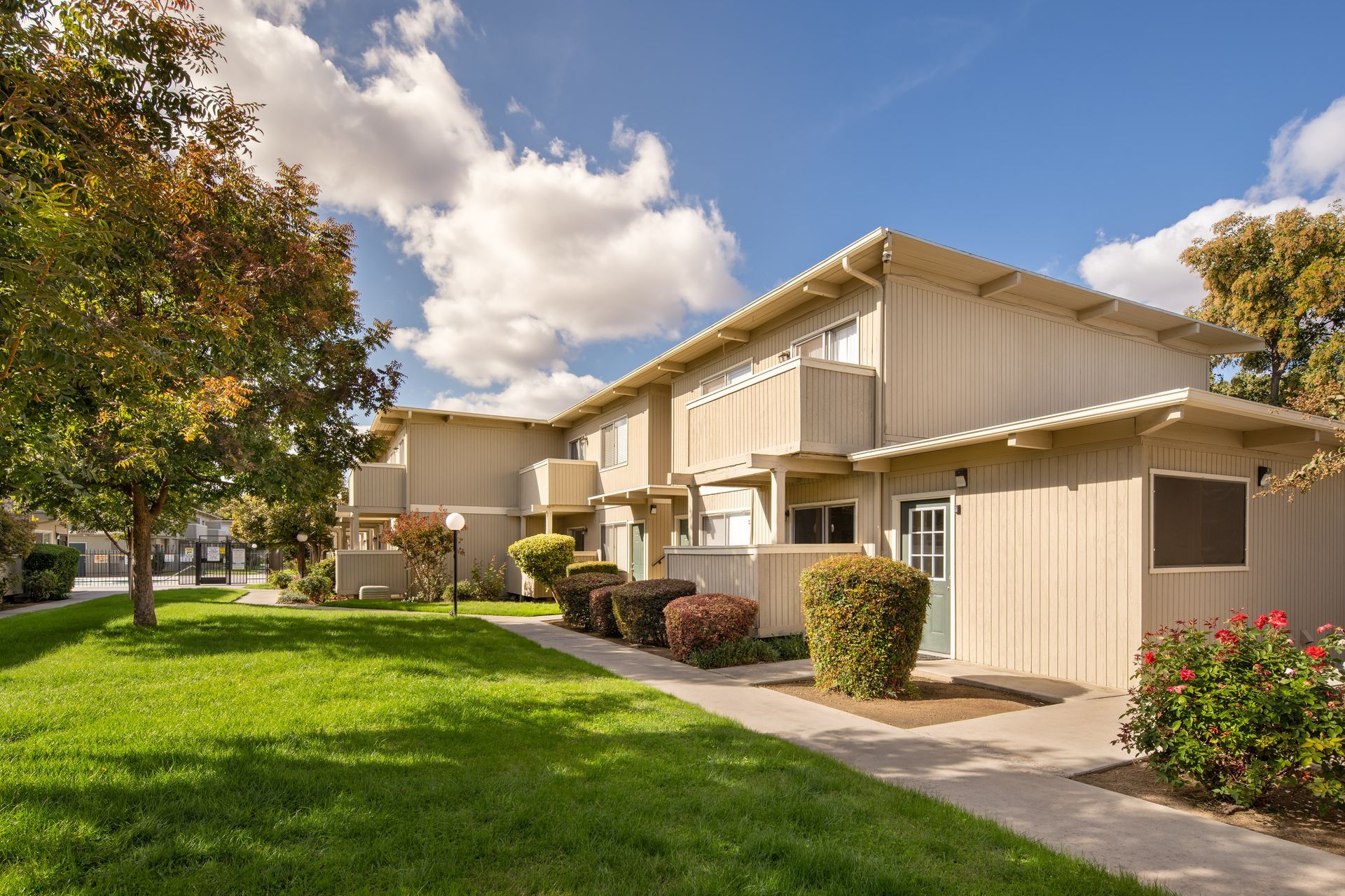 Two-story beige apartment building with balconies, green lawn, bushes, and a blue sky.