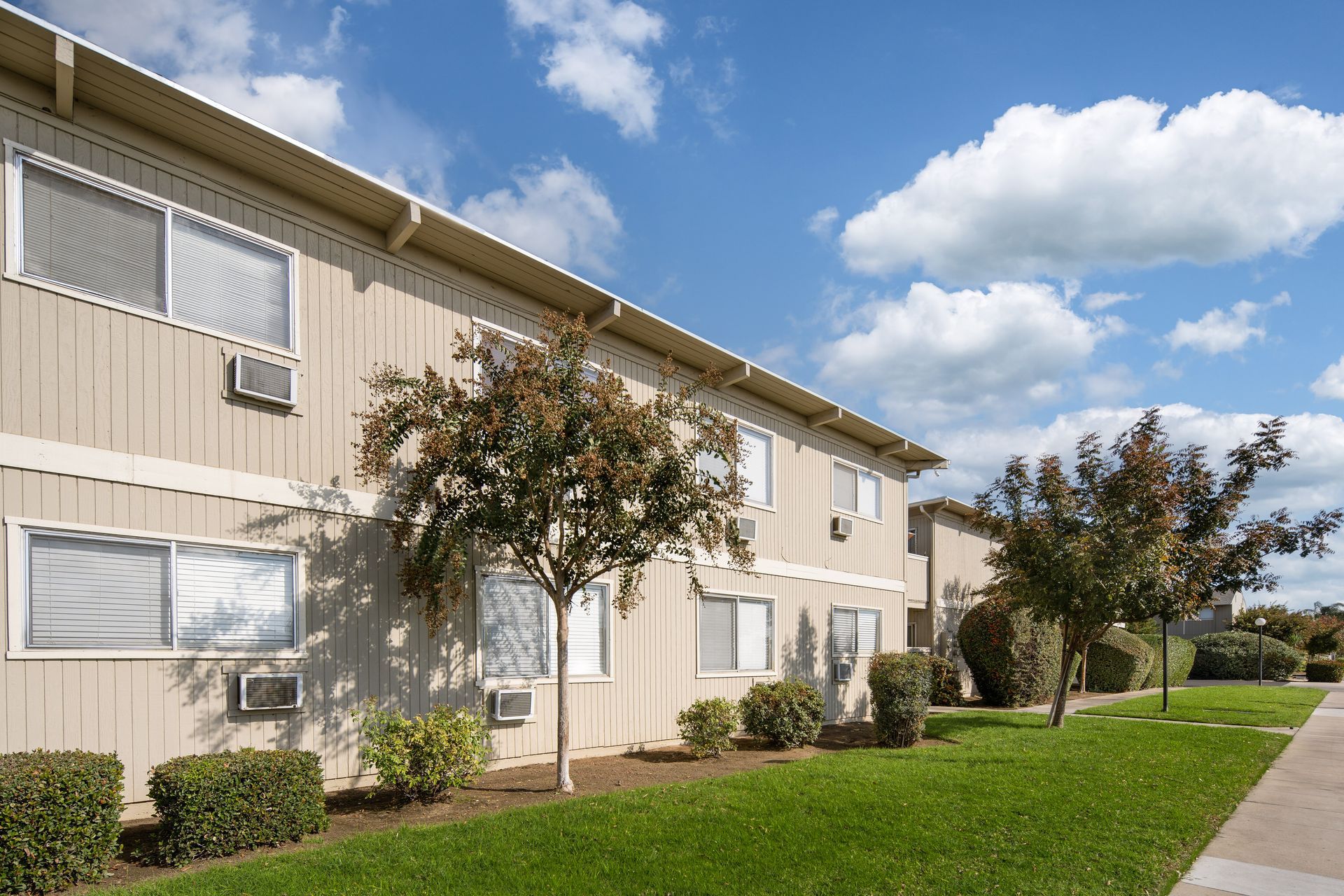 Apartment building with tan siding and green lawn under a blue sky with clouds.