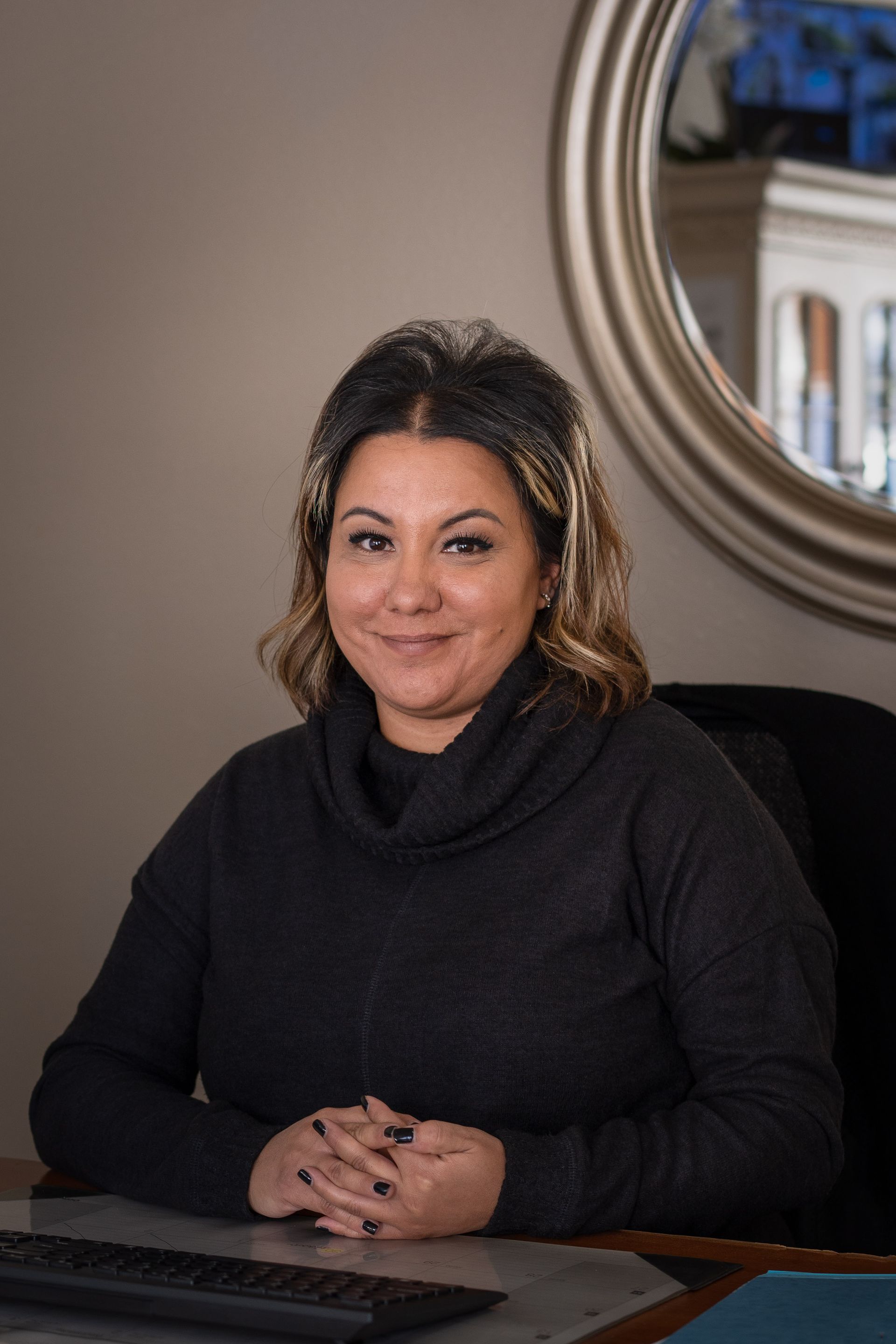 Woman sitting at a desk with folded hands, wearing a black sweater, smiling. A round mirror is in the background.