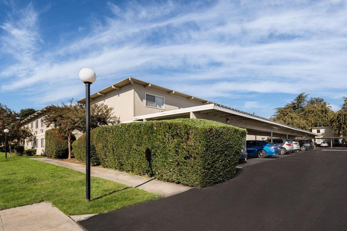 Apartment building with carport, parked cars, and manicured landscaping under a blue sky.