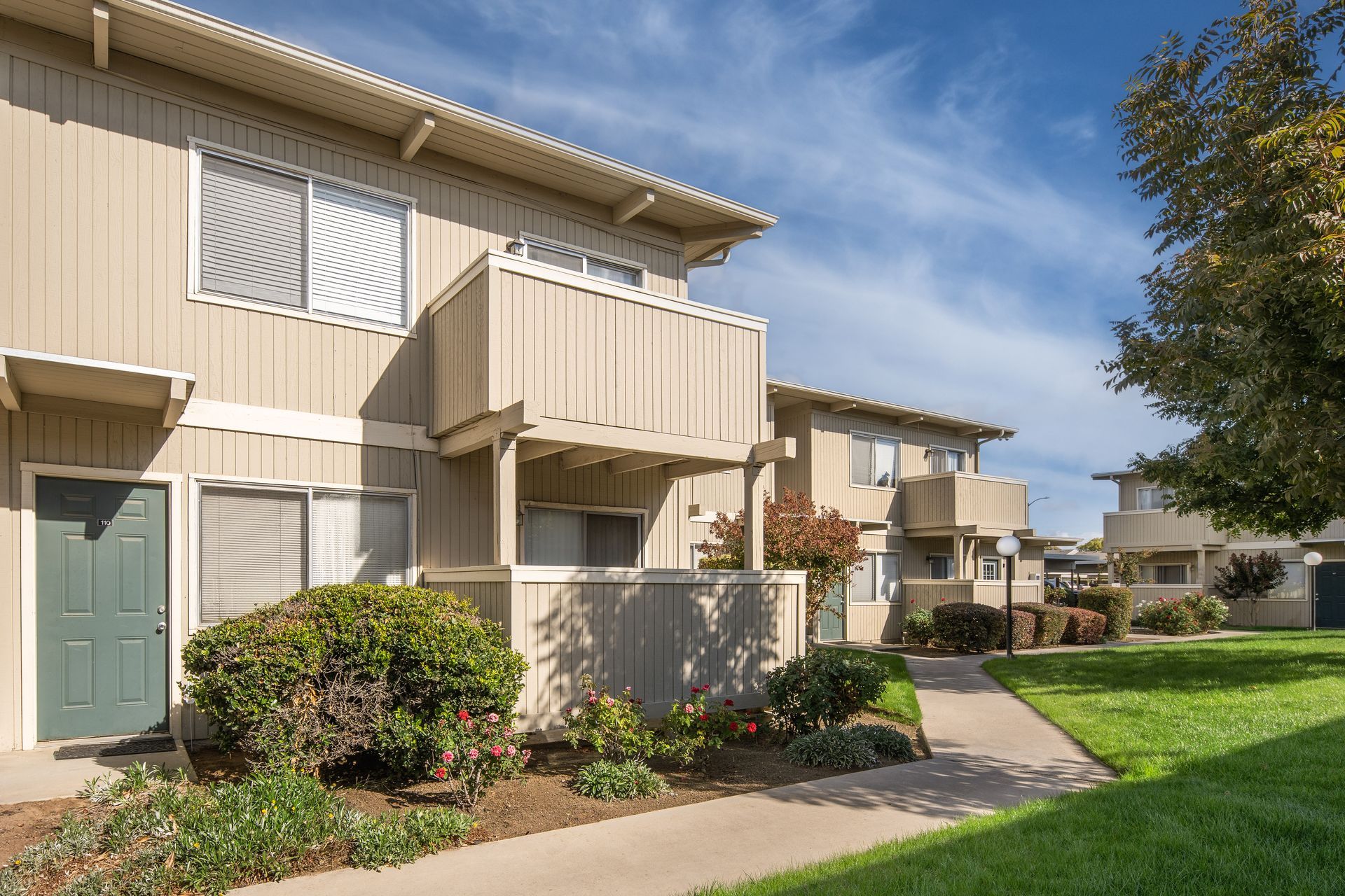 Apartment complex exterior with balconies, green door, beige siding, walkway, grass, and blue sky.