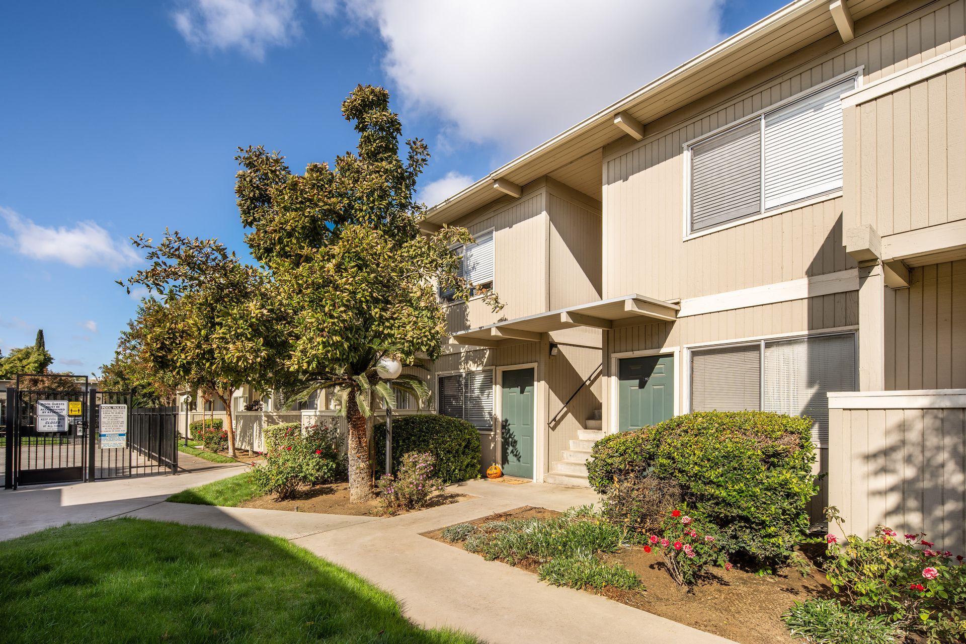 Apartment complex with beige buildings, green doors, and lush landscaping under a blue sky.