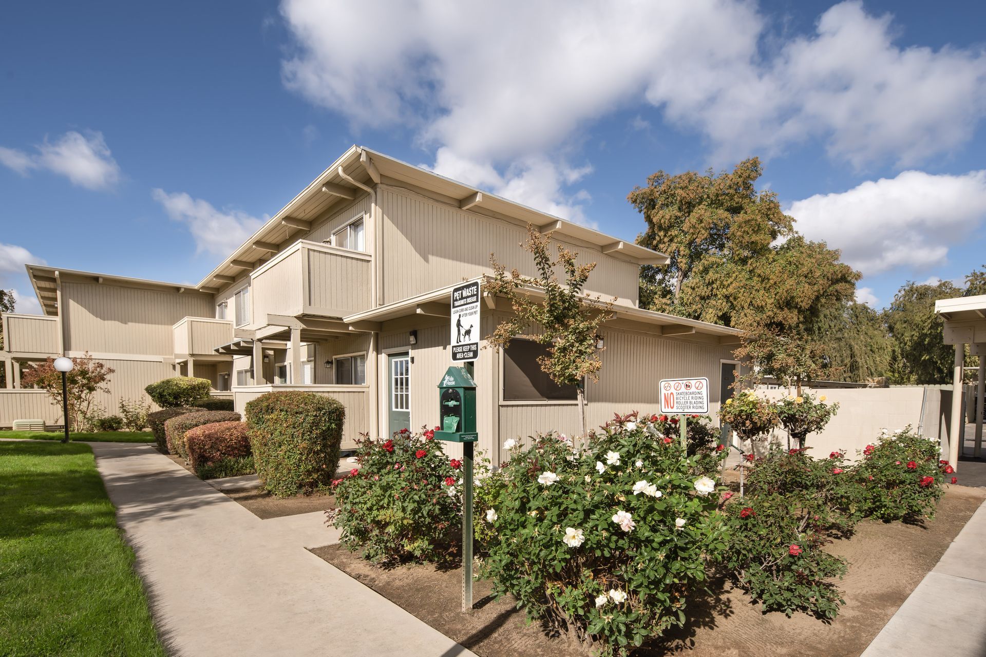 Apartment building with beige siding, a sidewalk, and rose bushes on a sunny day.