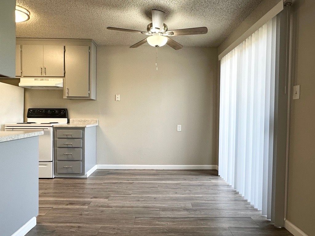 Kitchen and dining area with cabinets, stove, and sliding glass door with sheer curtains. Wooden floors.