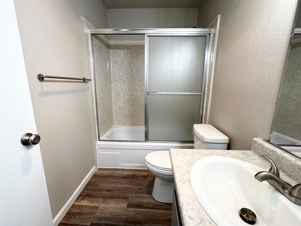 Bathroom with a white toilet, sink, and tub with sliding glass doors; wood-look floor.