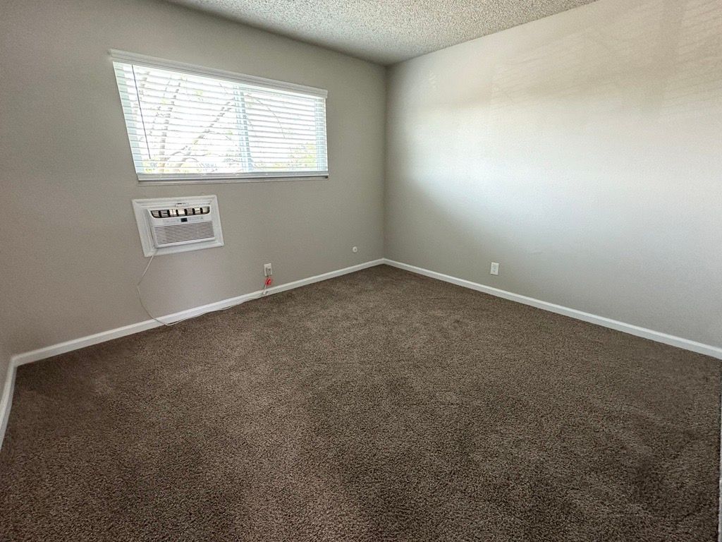 Empty room with brown carpet, window, and air conditioner unit. Gray walls and a textured ceiling.