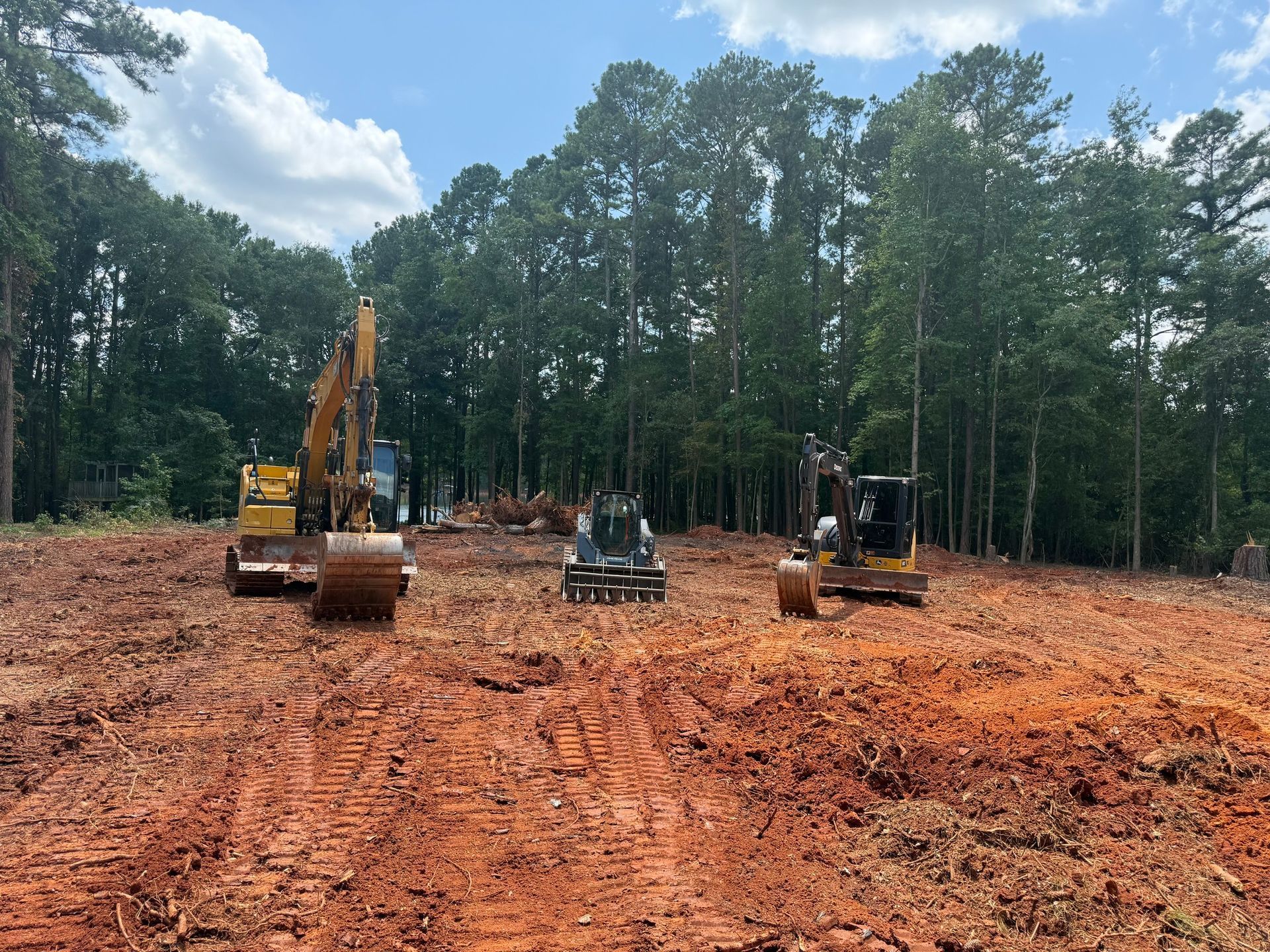Construction site with three excavators on red soil, trees in the background, blue sky with clouds.