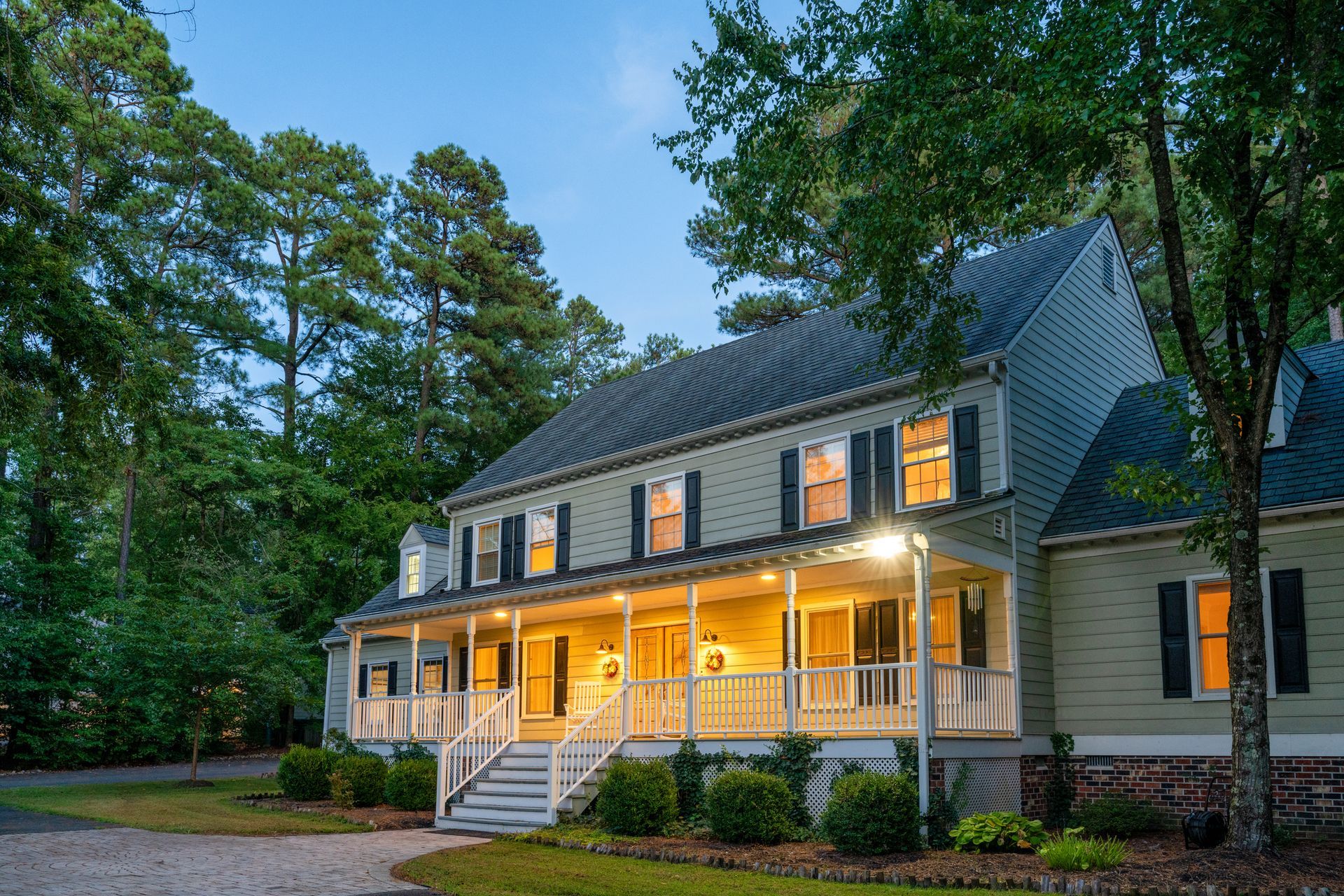 Two-story house with front porch lit up, green siding, surrounded by trees, dusk setting.