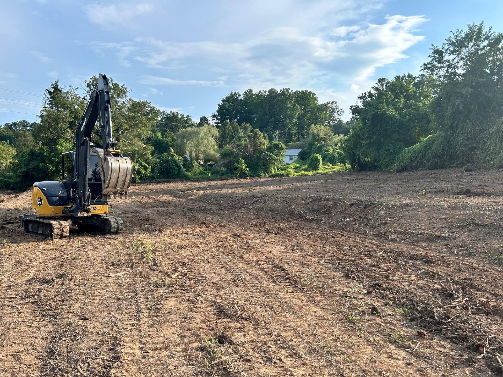 Excavator on cleared land, trees in background, cloudy sky.