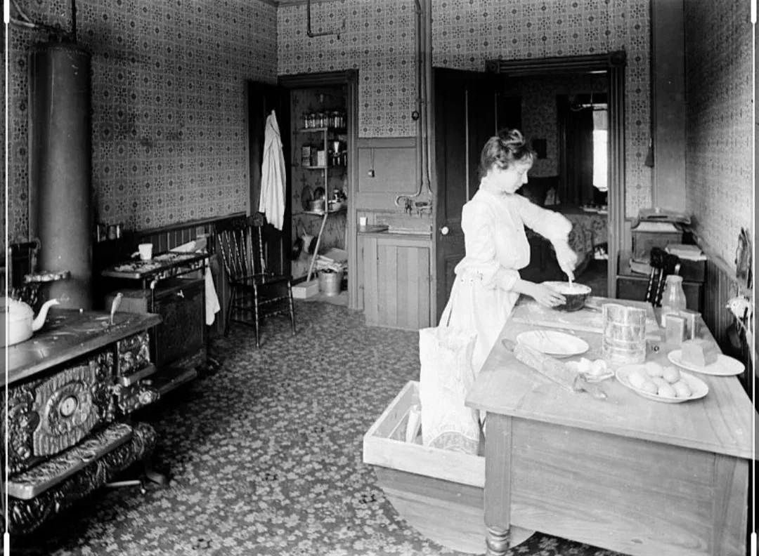 A black and white photo of a woman preparing food in a kitchen.