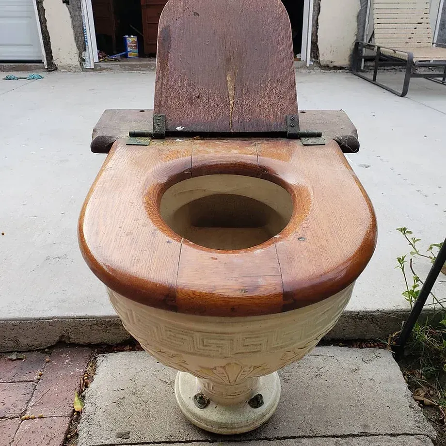 A toilet with a wooden seat sits outside in front of a garage