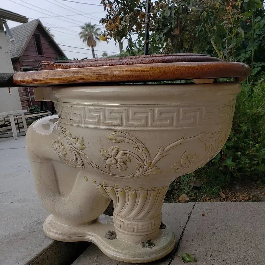 A white toilet with a wooden seat is sitting on a sidewalk.