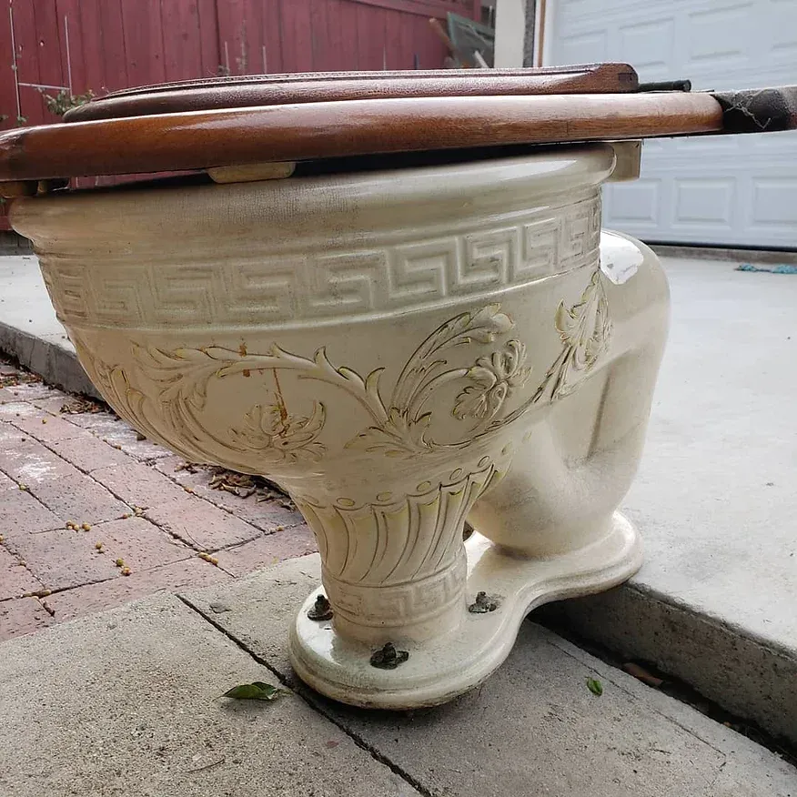 A white toilet with a wooden seat sits on a sidewalk