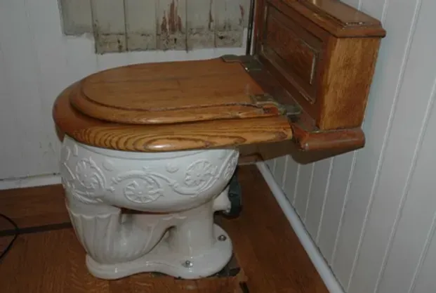 A toilet with a wooden seat is in a bathroom next to a window.