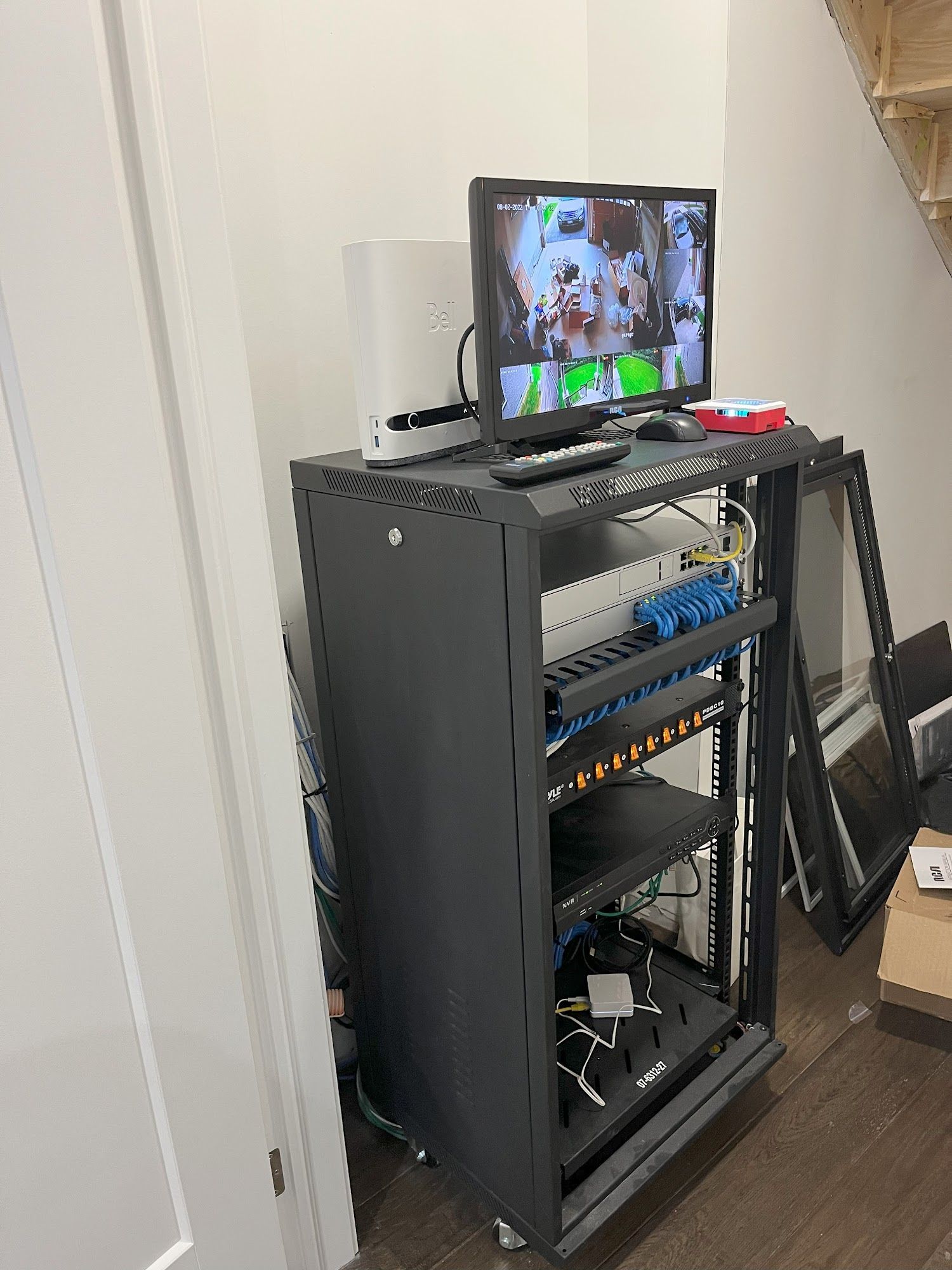 Black server rack with a monitor displaying security camera footage. Located near a white wall and stairs.