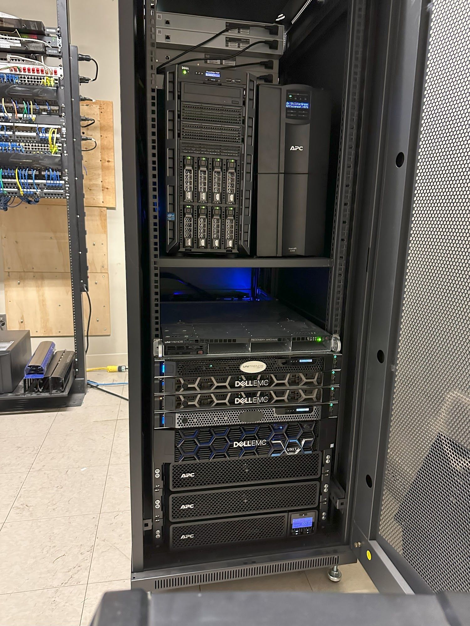 Server rack with black hardware, blue lights, and a power backup in a room.