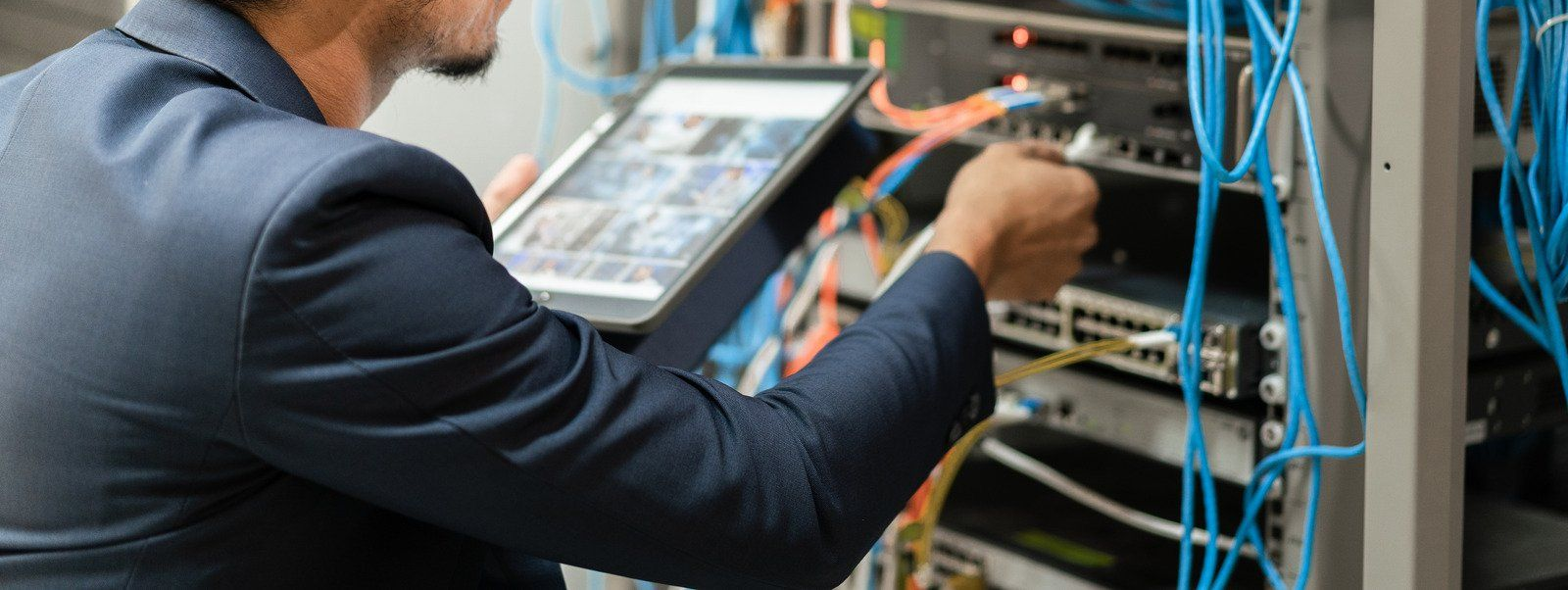 Man in suit working on server equipment, holding tablet. Blue and orange cables in server room.