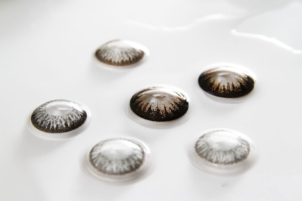 Contact lenses in various shades of gray and brown, resting in liquid on a white surface.