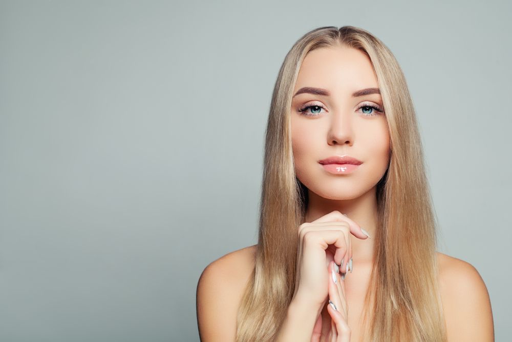 Blonde woman with straight hair, holding her hand near her face, looking at camera.