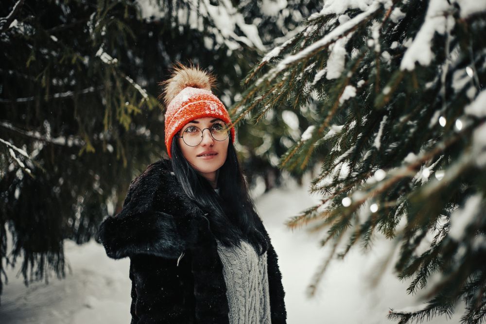 Woman in orange hat and black coat stands in snow-covered forest, looking towards the camera.