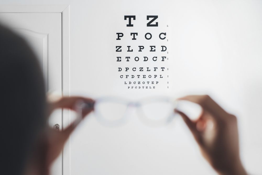 Person holding glasses in front of an eye chart. White wall, neutral setting.