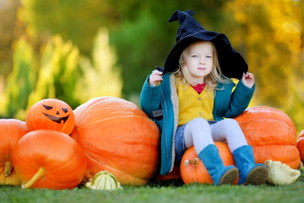 Girl in witch hat sits on pumpkins at a pumpkin patch.