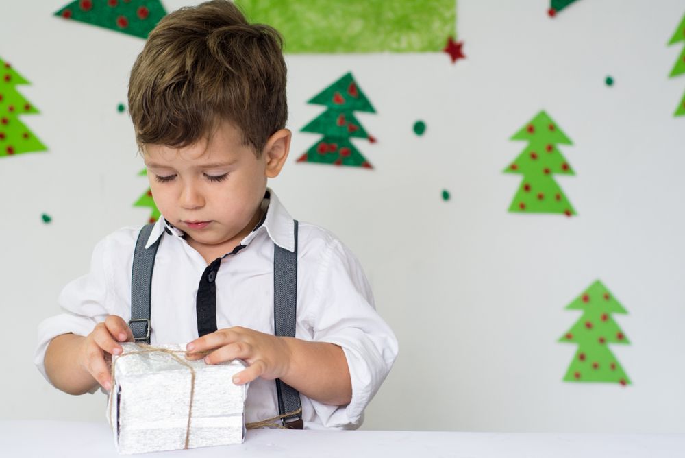 Boy in suspenders unwraps a small gift on a white surface, with Christmas tree decorations in the background.