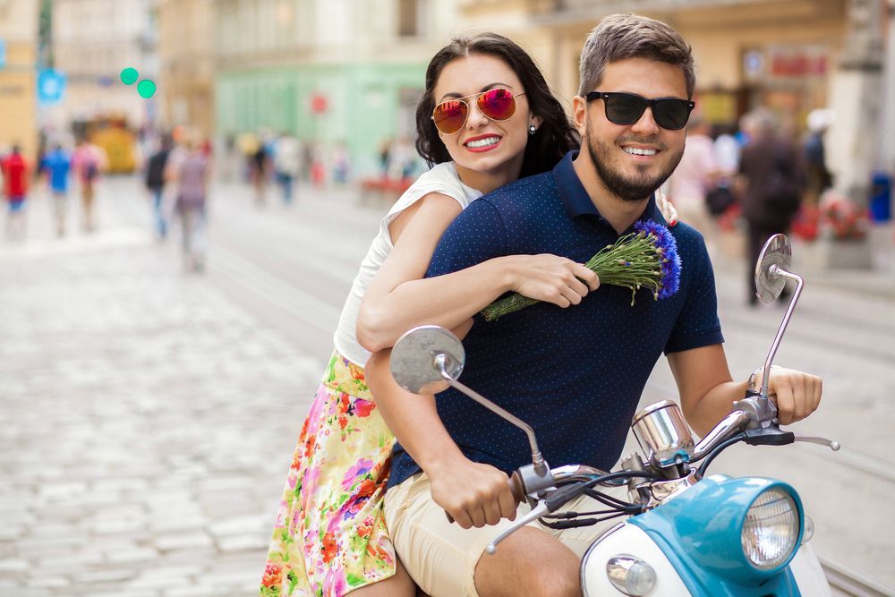 A man and a woman are wearing sunglasses and riding a scooter down a city street.